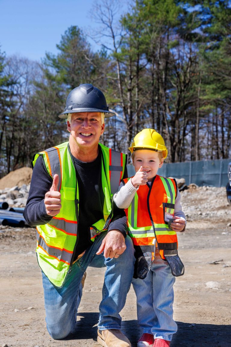 A worker in a hardhat with a young helper in a hardhat give thumbs up to the camera